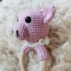 Vauvan virkattu helistin, vaaleanpunainen possun pää, valkoinen rusetti kaulassa. Puinen rengas