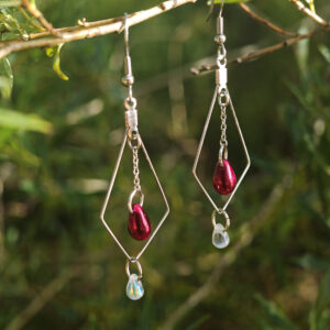 Hopean sävyiset korvakorut, jossa ruostumaton teräs kehikko, ketjussa punainen pisarahelmi ja alapuolella kirkas pisarahelmi.